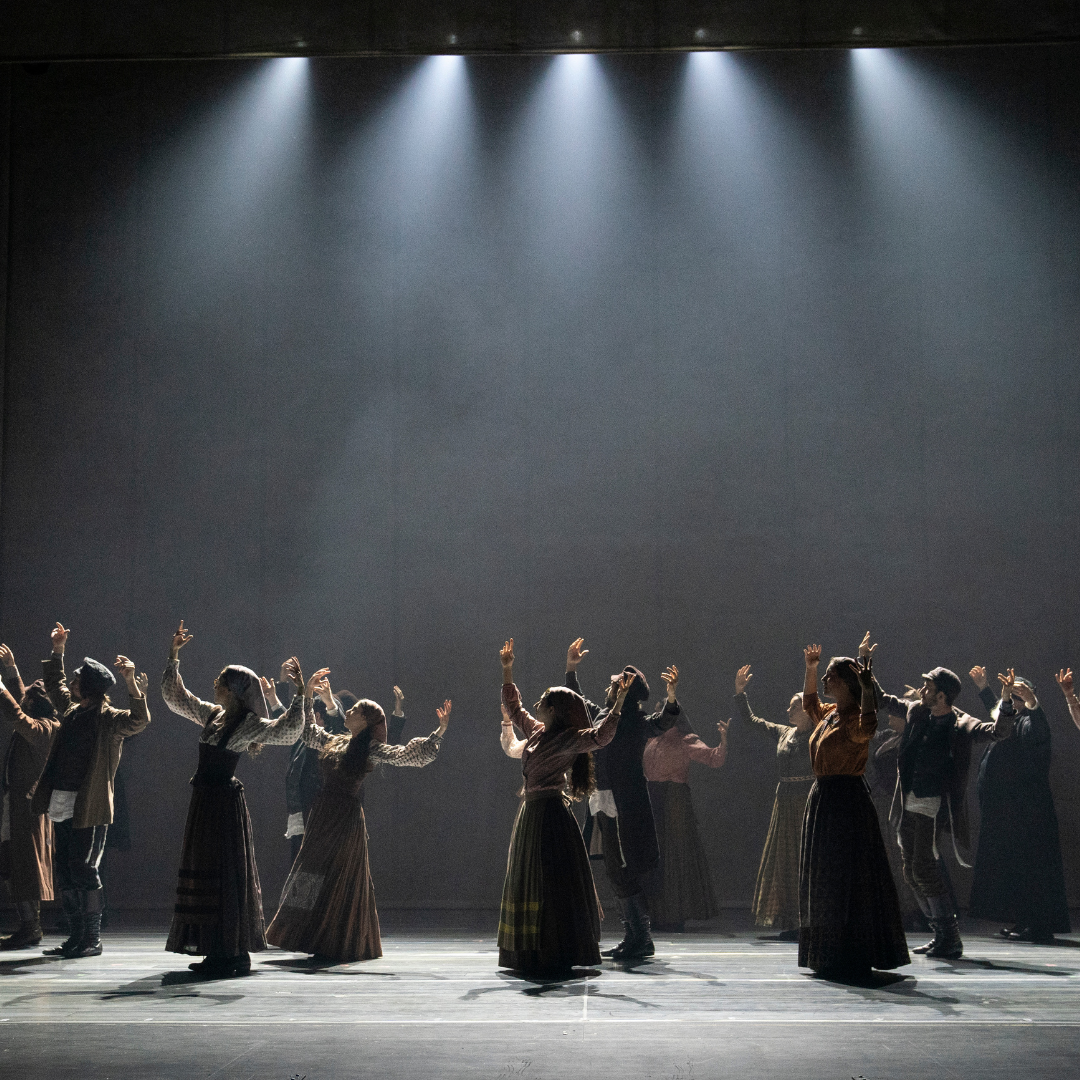 A dark stage with spotlights shining down on the cast of Fiddler on the roof, who are dancing in long skirts with their hands raised in the air.