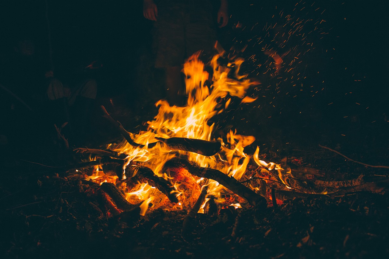 A campfire, glowing yellow and orange and burning atop a pile of woods against a dark background.