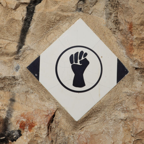 Image of a black painted fist in a circle on a white diamond on a stone wall.
