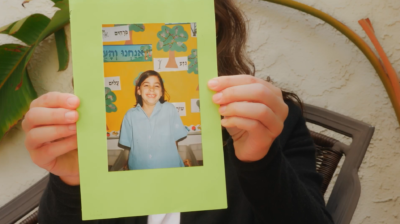 Simone Zimmerman shares a photo of herself as a child at Jewish Day School. The photo has a green frame, with a young child wearing a blue shirt smiling in front of a wall of Hebrew words. 